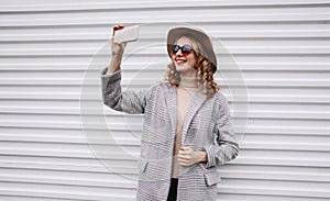 Stylish happy smiling woman taking selfie picture by smartphone in gray coat, round hat posing over white wall