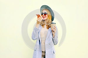 Stylish happy smiling young woman with smartphone using voice command recorder or calling, wearing coat jacket, round hat