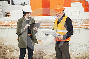 Stylish happy woman architect with tablet and senior foreman checking blueprints at construction site. Engineer and construction