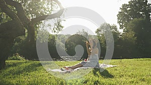 Stylish happy woman blowing bubbles in park by the river. Young brunette girl sitting on meadow near river, having fun, relaxing a