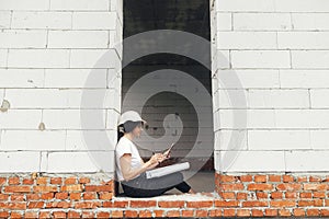 Stylish happy woman engineer in hard hat looking at digital plans on tablet while sitting in window of new modern house. Young