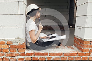 Stylish happy woman engineer in hard hat looking at digital plans on tablet while sitting in window of new modern house. Young