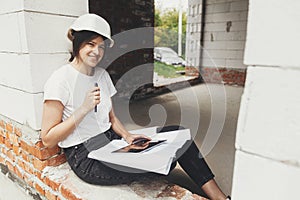 Stylish happy woman engineer in hard hat looking at digital plans on tablet while sitting in window of new modern house. Young