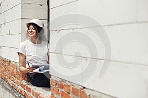 Stylish happy woman engineer in hard hat looking at digital plans on tablet while sitting in window of new modern house. Young