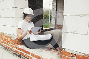 Stylish happy woman engineer in hard hat looking at digital plans on tablet while sitting in window of new modern house. Young