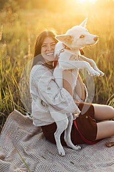 Stylish happy woman playing with her white dog on blanket in warm sunset light in summer meadow