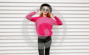 Stylish happy young woman in colorful pink knitted sweater, black round hat posing on city white wall