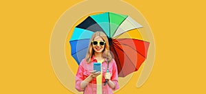 Stylish happy young woman looking at mobile phone holds colorful umbrella wearing pink jacket, sunglasses on yellow studio