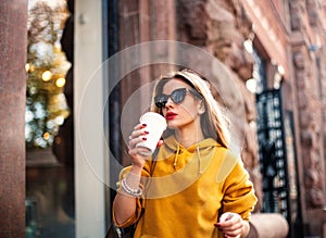 Stylish happy young woman wearing boyfrend jeans, white sneakers bright yellow sweatshirt.She holds coffee to go. portrait of smil