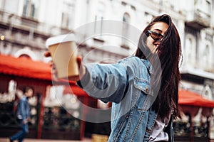Stylish happy young woman wearing woman in a denim jacket. She holds coffee to go. portrait of smiling girl in sunglasses and bag