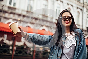 Stylish happy young woman wearing woman in a denim jacket. She holds coffee to go. portrait of smiling girl in sunglasses and bag