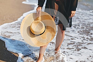 Stylish hat in hands of carefree woman walking in sea waves on sunny beach, close up. Summer vacation. Young fashionable female