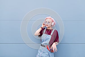 Stylish hipist girl with pink hair stands on the background of a blue wall and listens to music in the headphones