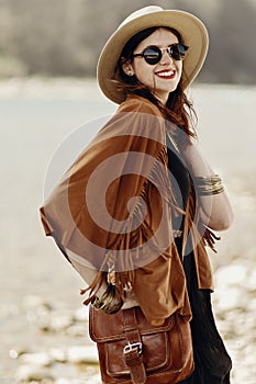 stylish hipster boho woman smiling in sunglasses with hat, leather bag, fringe poncho and accessory. happy traveler girl look, ne