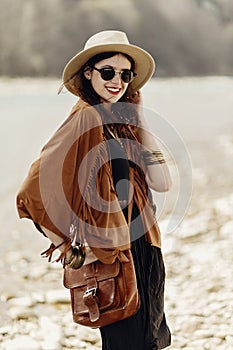 stylish hipster boho woman smiling in sunglasses with hat, leather bag, fringe poncho and accessory. traveler girl look, near