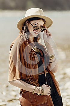 stylish hipster boho woman smiling in sunglasses with hat, leather bag, fringe poncho and accessory. traveler girl look, near