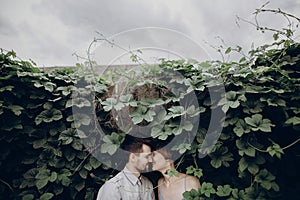 Stylish hipster bride and groom kissing in green leaves, holding