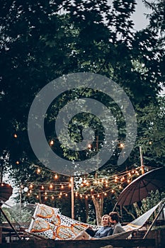 Stylish hipster couple cuddling and relaxing in hammock under retro lights in evening summer park. man hugging woman and resting