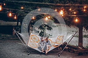 Stylish hipster couple cuddling and relaxing in hammock under retro lights in evening summer park. man hugging woman and resting