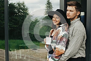Stylish hipster couple with morning coffee relaxing on background of modern cabin and big windows in mountains. Happy young family