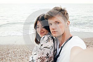 Stylish hipster couple taking selfie on beach at evening sea. Summer vacation. Portrait of happy young family on honeymoon on
