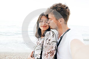 Stylish hipster couple taking selfie on beach and kissing at evening sea. Summer vacation. Portrait of happy young family on