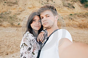 Stylish hipster couple taking selfie on beach at sandy cliff at sea. Summer vacation. Portrait of happy young family on honeymoon