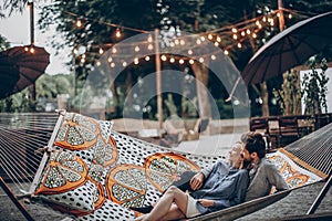 Stylish hipster family cuddling and relaxing in hammock under retro lights in evening summer park. rustic man and woman embracing