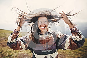 Stylish hipster girl with backpack and windy hair smiling on top of mountains. Portrait of happy young woman relaxing. Carefree