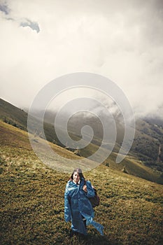 Stylish hipster girl in blue raincoat and with backpack standing on top of misty mountains and clouds. Young woman traveler