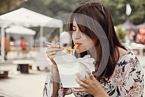 Stylish hipster girl eating wok noodles with vegetables from carton box with bamboo chopsticks. Asian Street food festival. Happy