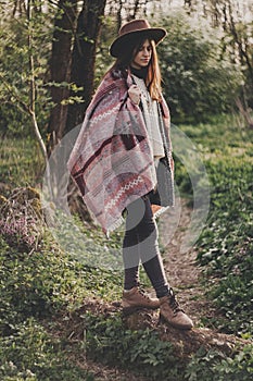 stylish hipster girl in hat and poncho posing in woods in evening sunshine. bohemian fashionable woman traveler in sunlight . spa