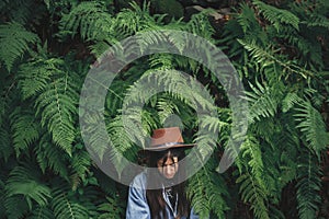 Stylish hipster girl in hat posing in big fern leaves in forest. Portrait of calm young woman in green leaves in woods. Travel and