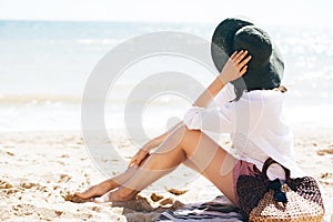 Stylish hipster girl in hat sitting on beach with straw bag and tanning near sea waves. Summer vacation. Happy boho woman relaxing