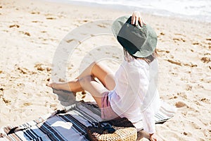 Stylish hipster girl in hat sitting on beach with straw bag and tanning. Summer vacation. Happy young boho woman relaxing and