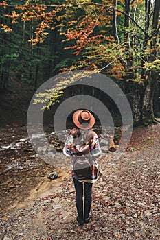 Stylish hipster girl in hat traveling in mountains. Young woman with backpack exploring and walking in summer forest. Travel and