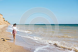 Stylish hipster girl in hat walking on beach and smiling. Summer vacation. Happy young boho woman relaxing and enjoying sunny warm