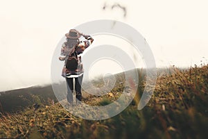 Stylish hipster girl in hat walking on top of mountains. Happy young woman with backpack exploring misty mountains. Travel and
