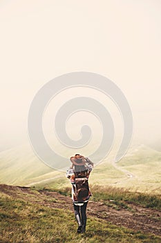 Stylish hipster girl in hat walking on top of mountains. Happy young woman with backpack exploring misty mountains. Travel and