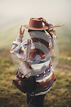 Stylish hipster girl in hat walking on top of mountains. Happy young woman with backpack exploring misty mountains. Travel and