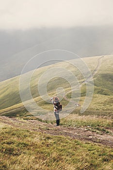 Stylish hipster girl in hat walking on top of mountains. Happy young woman with backpack exploring misty mountains. Travel and