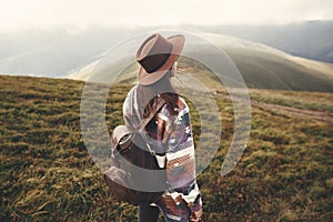 Stylish hipster girl in hat walking on top of mountains. Happy young woman with backpack exploring sunny mountains. Travel and