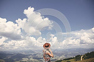 Stylish hipster girl in hat walking on top of sunny mountains. Happy young woman exploring in summer mountains and looking at sky