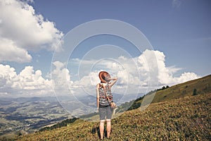 Stylish hipster girl in hat walking on top of sunny mountains. Happy young woman exploring in summer mountains and looking at sky