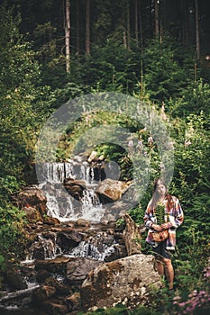 Stylish hipster girl holding fern leaf and relaxing in forest in mountains. Young woman traveler exploring waterfall woods. Eco