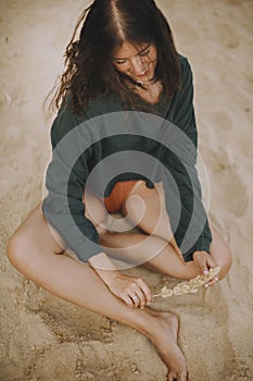 Stylish hipster girl holding herb and sitting on sandy beach. Fashionable tanned young woman in modern swimsuit and sweater