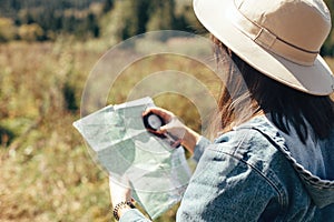 Stylish hipster girl holding map and compass, traveling in sunny mountains. Woman in hat exploring map and hiking on top of