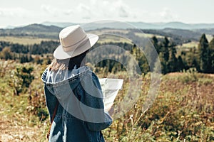 Stylish hipster girl holding map and traveling in sunny mountains. Woman in hat exploring map and hiking on top of mountain on