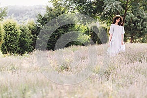 Stylish hipster girl in linen dress and hat relaxing in lavender field near tree, focus on lavender. Bohemian woman enjoying