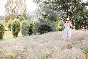 Stylish hipster girl in linen dress and hat relaxing in lavender field near tree, focus on lavender. Bohemian woman enjoying